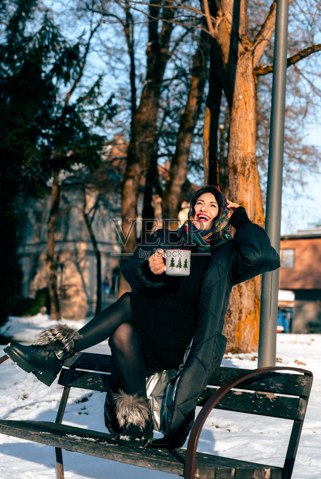 身着冬装的女士微笑着坐在公园长椅上，手中拿着咖啡杯，周围是覆盖着雪的树木，她面带喜悦地享受着户外的晴朗天气照片摄影图片