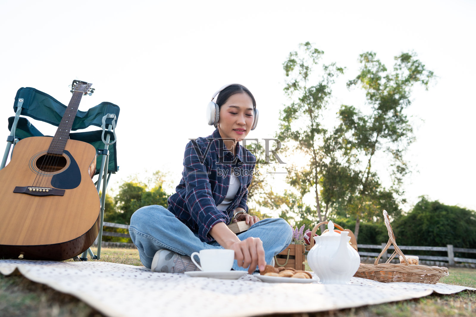 夕阳下，一位戴着耳机、吃着饼干、拿着咖啡杯的快乐年轻亚洲女性，在公园的野餐毯上弹着木吉他放松。照片摄影图片