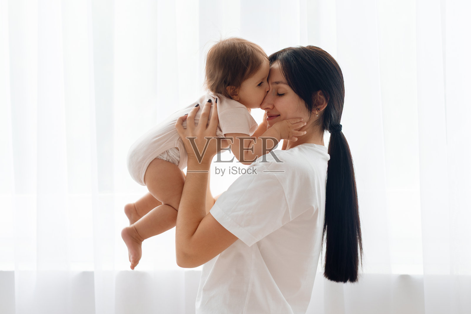 在柔和光线照亮的室内，一位母亲在家中亲吻她的婴儿照片摄影图片