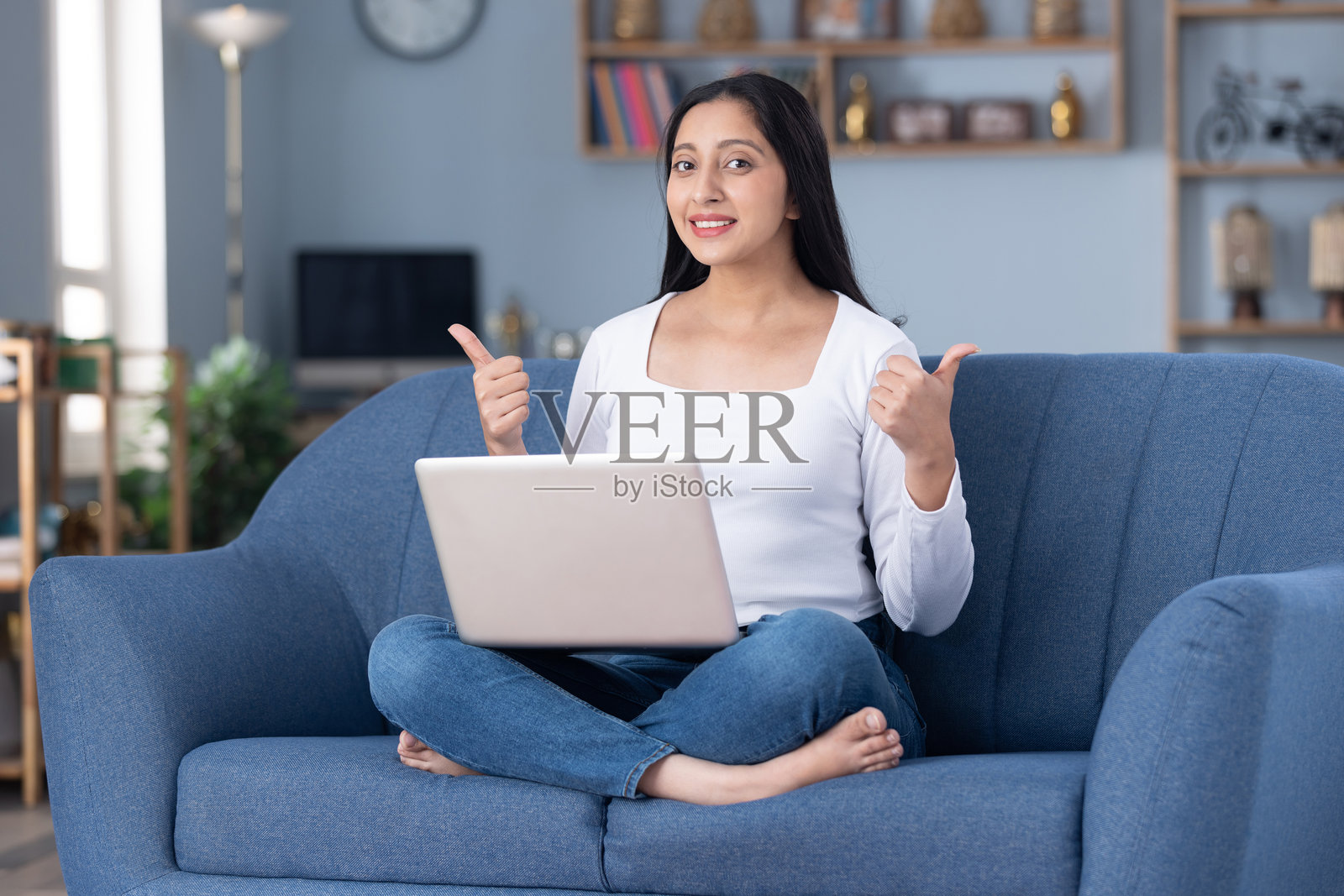 照片：穿着时尚衣服的漂亮年轻女孩在家里的沙发上使用笔记本电脑照片摄影图片