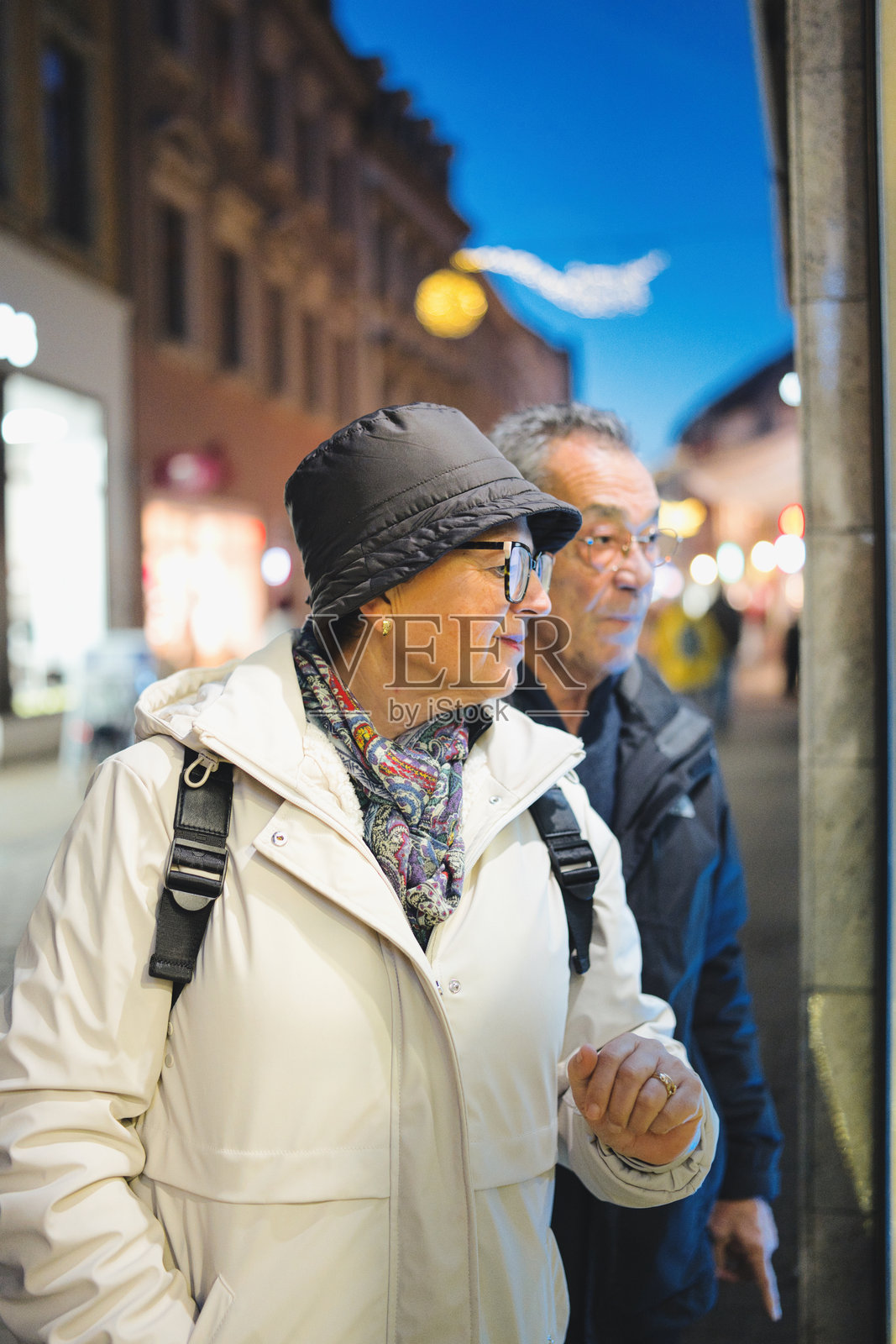 傍晚时分，一对成熟的夫妇在欧洲城市街道上逛街，走过亮着灯的橱窗展示，他们穿着夹克、围着围巾，享受着夜晚的出行。照片摄影图片