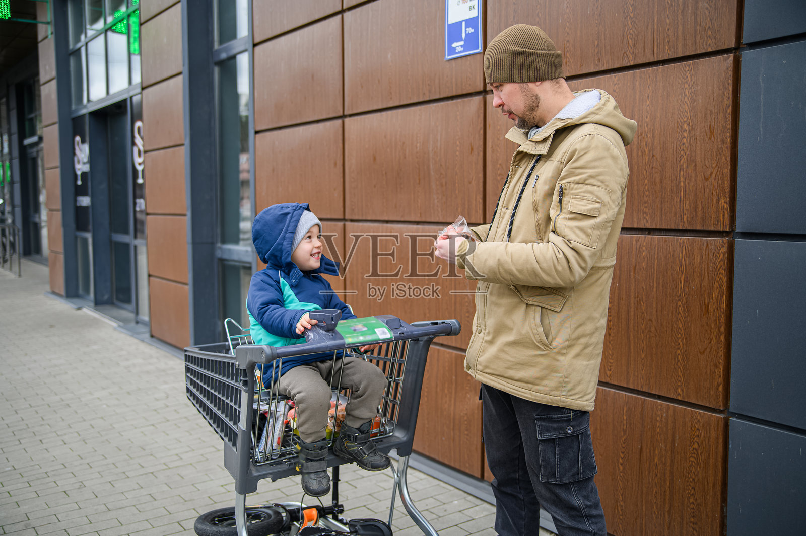父亲和儿子在冬季的超市购物之旅中共享天伦之乐，他们推着购物车，分享零食，并在商店外享受着平凡的家庭时光。照片摄影图片