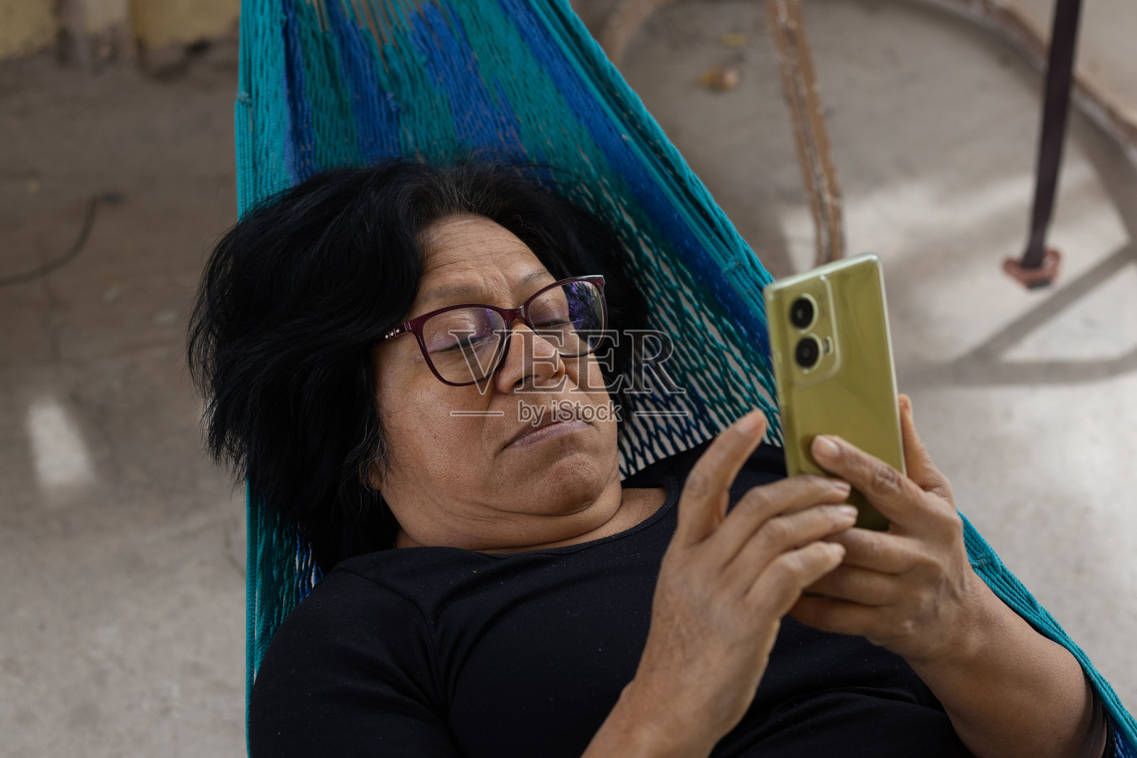 一位女士戴着眼镜，舒适地躺在色彩鲜艳的吊床上，一边看手机一边享受户外的悠闲时光，尽情放松。照片摄影图片