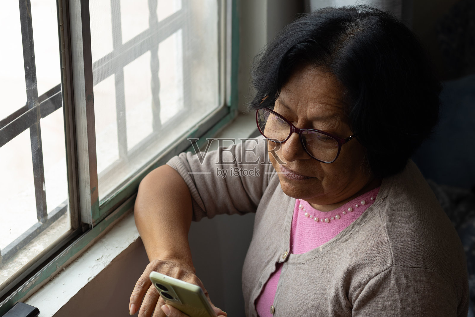 一位年长的西班牙裔女性在室内窗户边，手持智能手机，与科技互动，与他人建立联系和沟通照片摄影图片