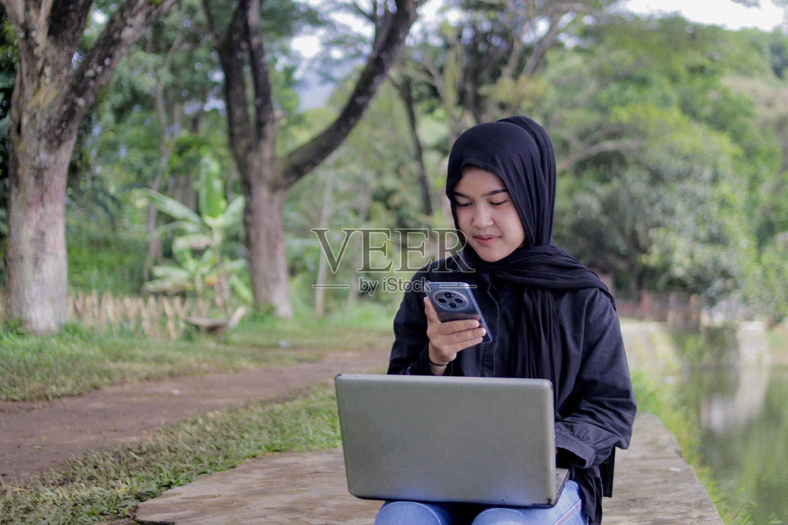 年轻的亚洲女性戴着头巾，在户外使用笔记本电脑工作时使用智能手机照片摄影图片