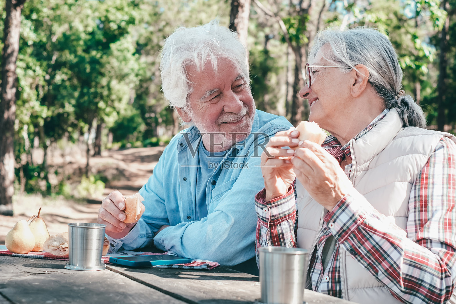 一对快乐的老年夫妇在阳光明媚的森林里享受户外野餐。这位年长的男士和女士坐在一张木桌旁，微笑着分享食物和饮料，展现了积极的退休自然生活方式。照片摄影图片