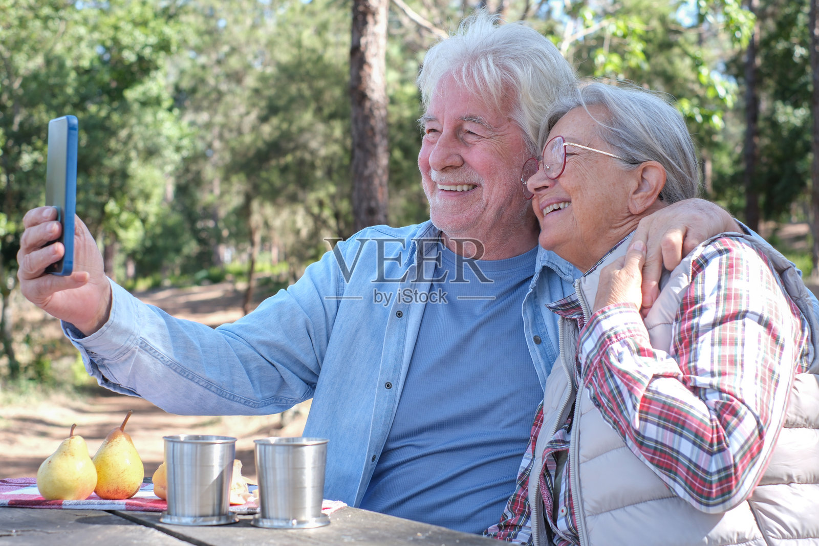 一对快乐的老年夫妇在阳光明媚的森林里享受户外野餐，并使用智能手机。年长的夫妇坐在木桌旁，微笑着分享食物和饮料。积极的退休生活在大自然中。照片摄影图片