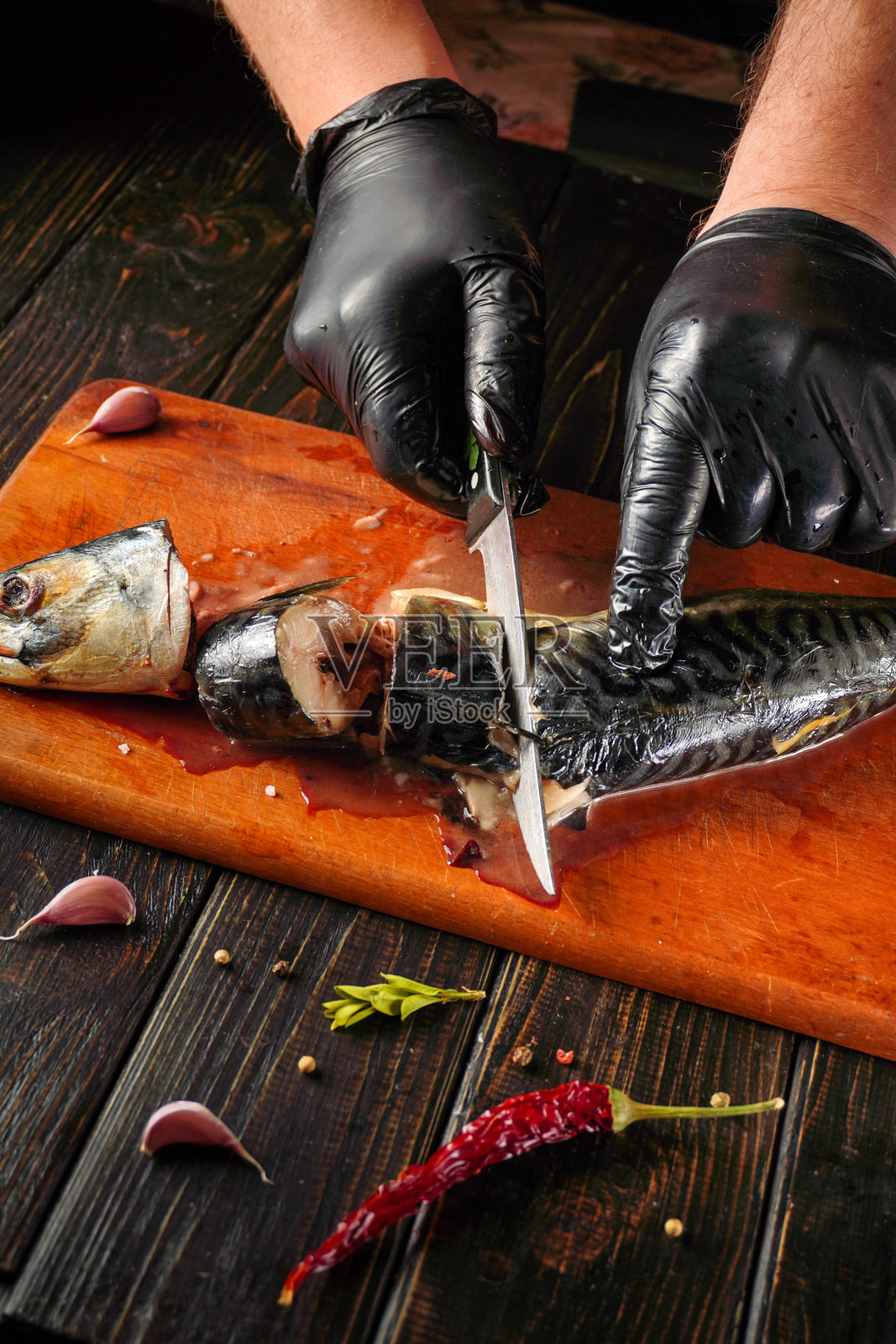一个人正用刀在木质表面上切割鲭鱼。旁边有大蒜、胡椒和辣椒。为保持卫生，手上戴着手套。照片摄影图片