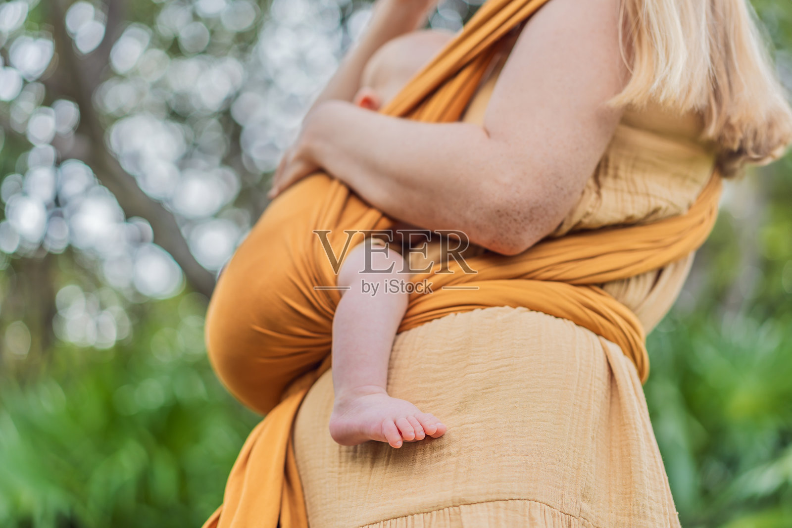 一位母亲在公园里，用黄色的背带抱着她的小宝贝。温馨有爱的家庭时刻。亲子背带、育儿、母子情深的理念照片摄影图片