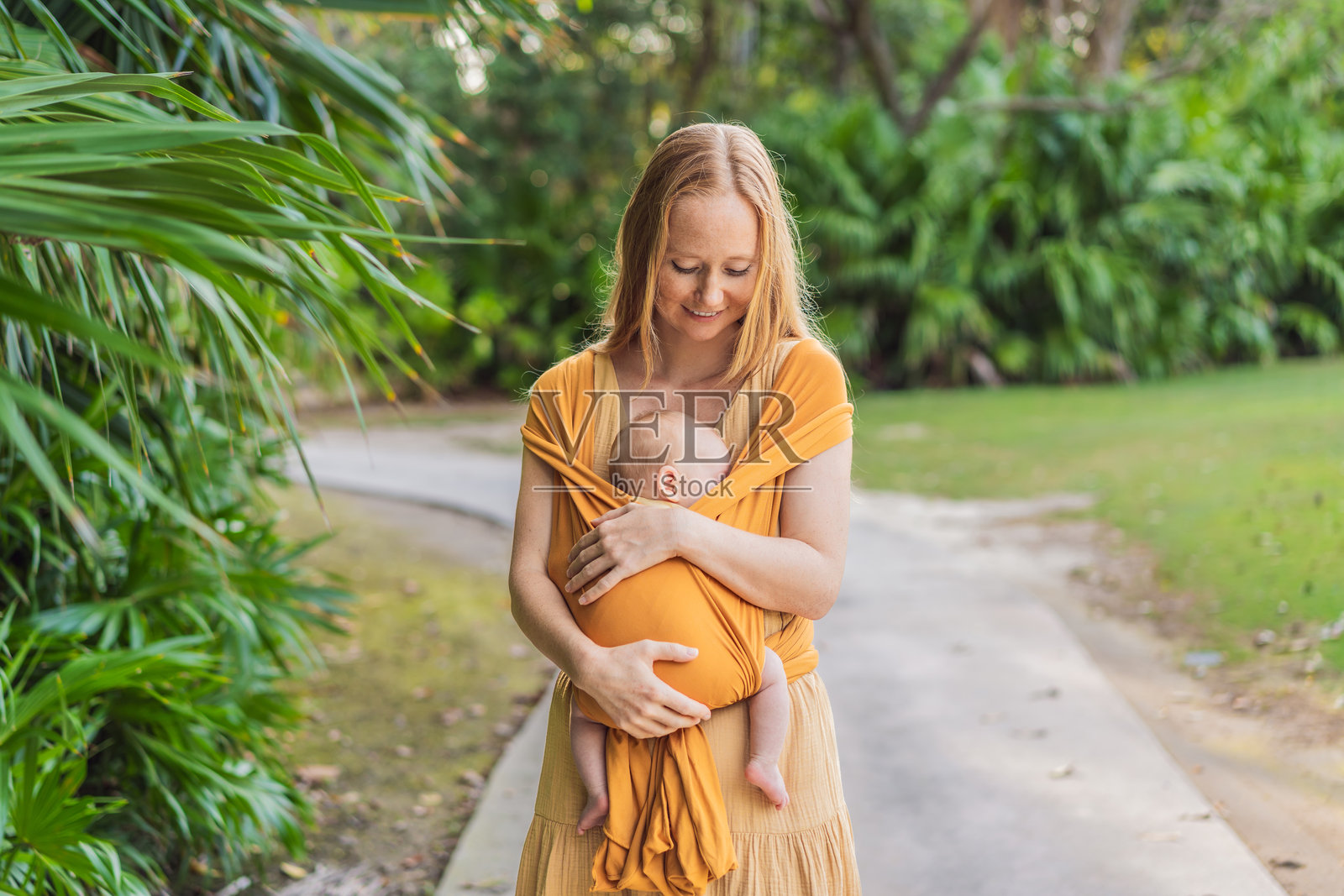 妈妈在公园里用黄色的背带抱着她的小宝贝。温馨有爱的家庭时刻。亲子背带、育儿和母子情深的理念照片摄影图片