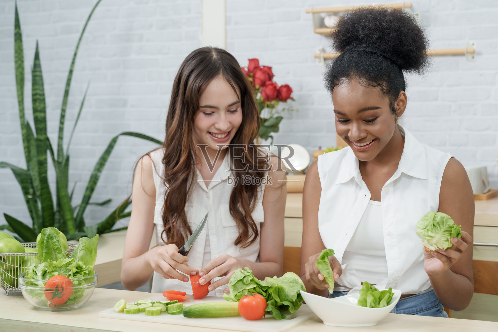 厨房里，不同种族、笑容灿烂的少女们正在一起准备健康食物。一位亚洲女孩在切番茄，她非裔朋友则在准备生菜。这个场景展现了美丽的肌肤和健康的生活方式。照片摄影图片
