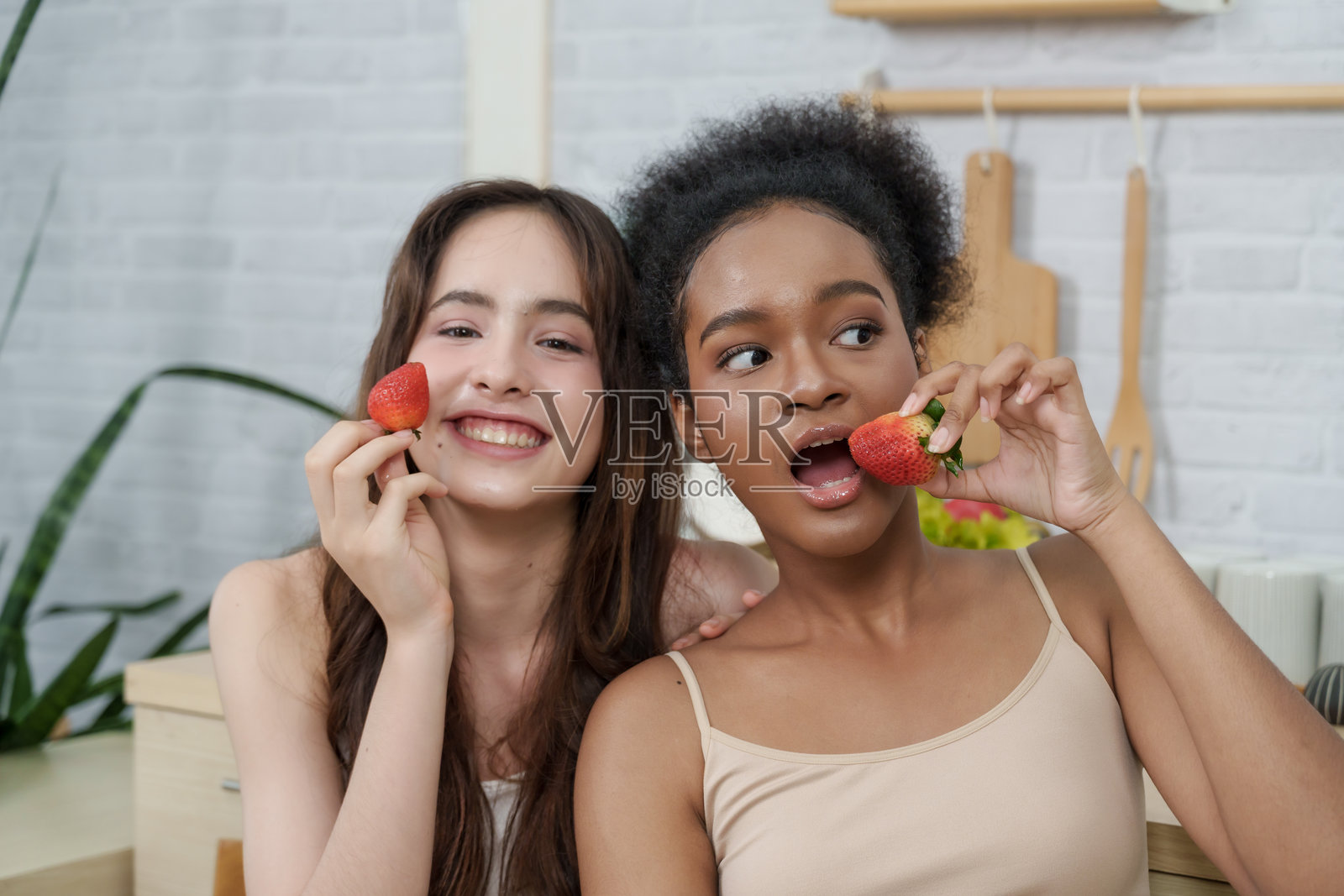 两位年轻的亚裔、白人和非裔女性在家庭厨房里享用新鲜草莓零食，分享着玩闹的时刻，体现了健康饮食理念、生活方式、友谊、天然食物选择以及社交联系。照片摄影图片