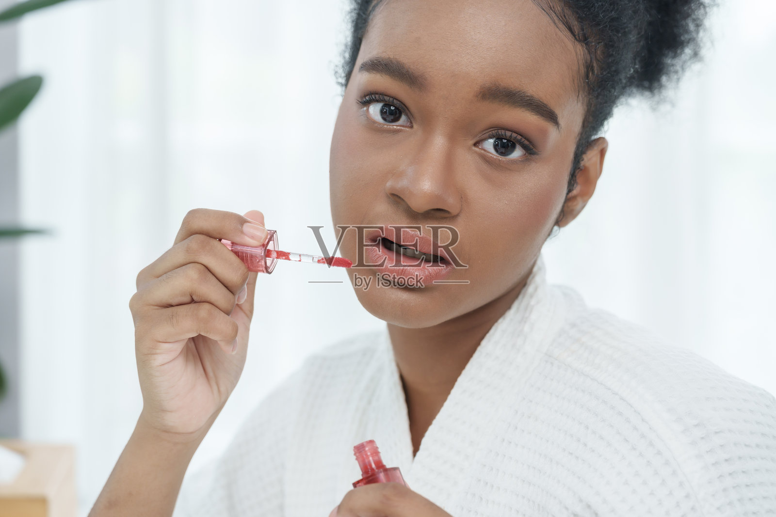 特写镜头：一位非洲少女在使用唇彩棒，在涂抹化妆品前，嘴唇微张，她穿着浴袍，在柔和的室内光线下，表情平静，正在进行早晨的美容护理。照片摄影图片