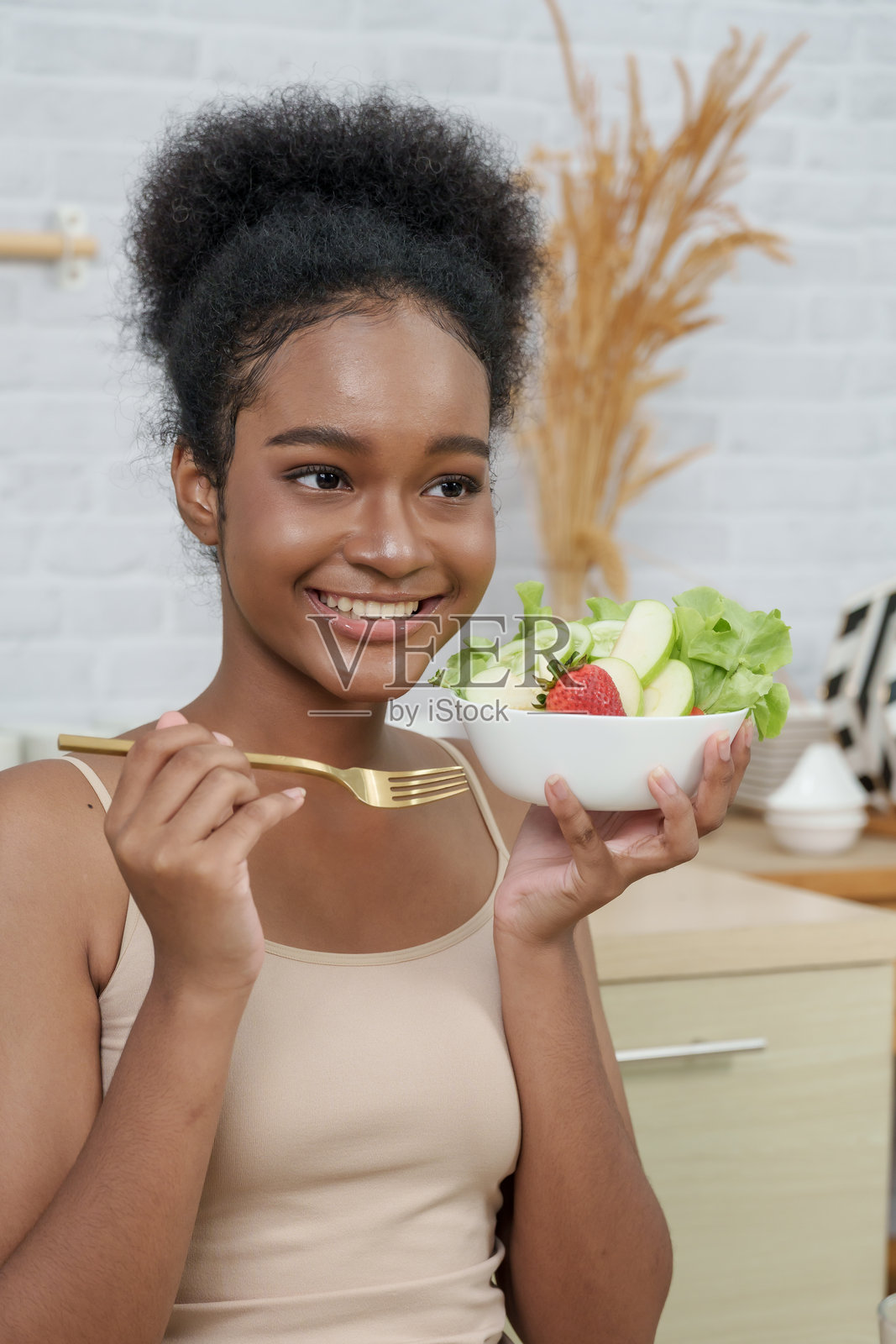 年轻的多民族女性在现代厨房里享用一碗健康沙拉，她微笑着，手里拿着叉子，通过营养的食物选择来代表自我关怀、美丽和容光焕发的皮肤的概念照片摄影图片