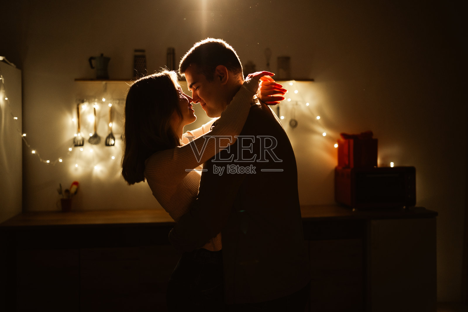 夜色厨房里，一对恋人拥抱的剪影，背景是闪烁的灯串。浪漫时刻，爱与激情。照片摄影图片
