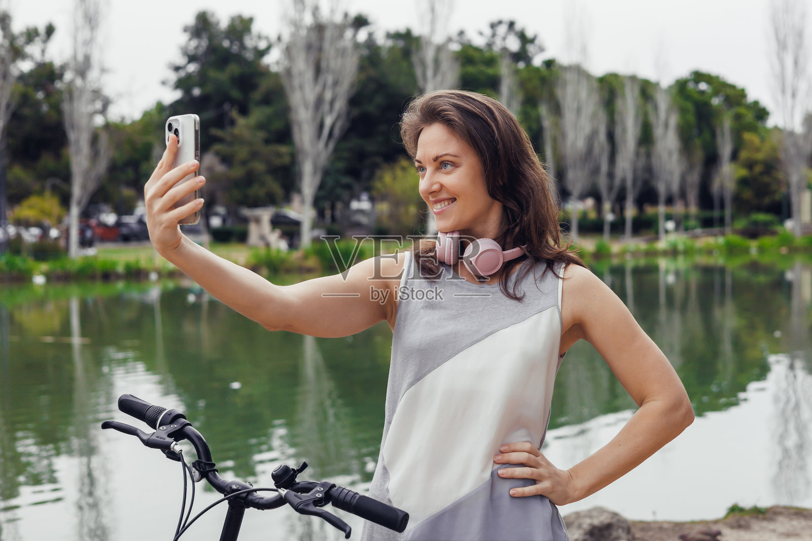 一位棕色头发的年轻女子戴着耳机微笑着，她将智能手机靠近一个被绿植环绕的宁静池塘。照片摄影图片
