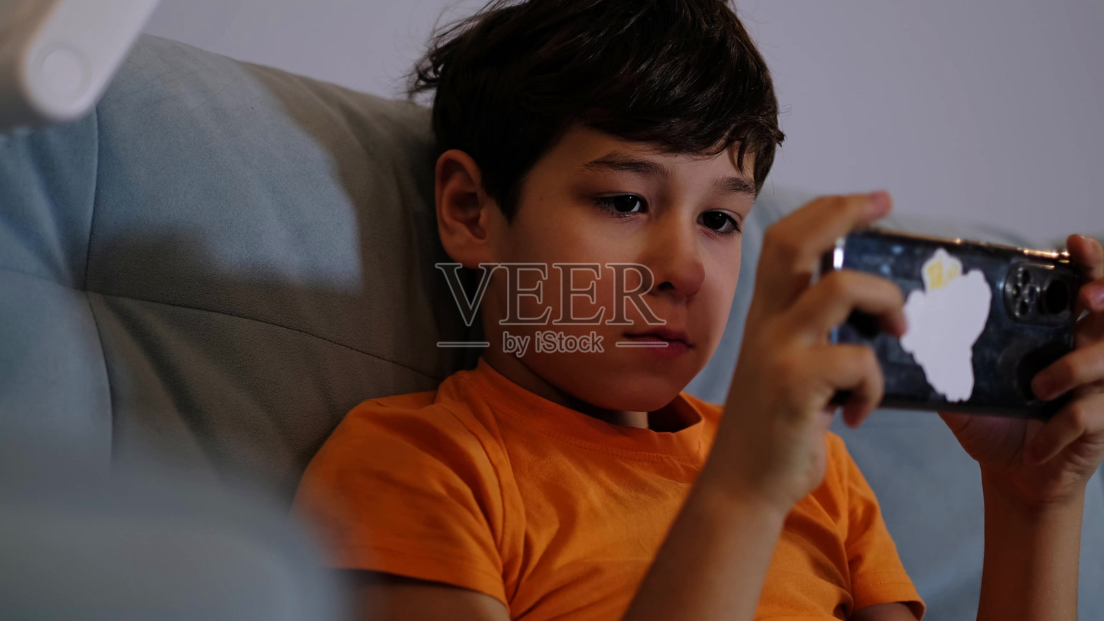 一个穿着橙色衬衫的男孩坐在沙发上，兴奋地玩着手机游戏。这个快乐的时刻突显了科技在童年中的吸引力。照片摄影图片