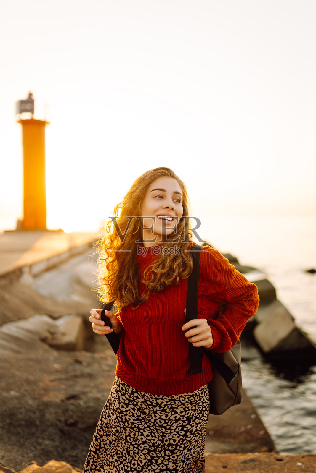 一位背着背包的年轻女子正走向灯塔，欣赏着日落美景。照片摄影图片