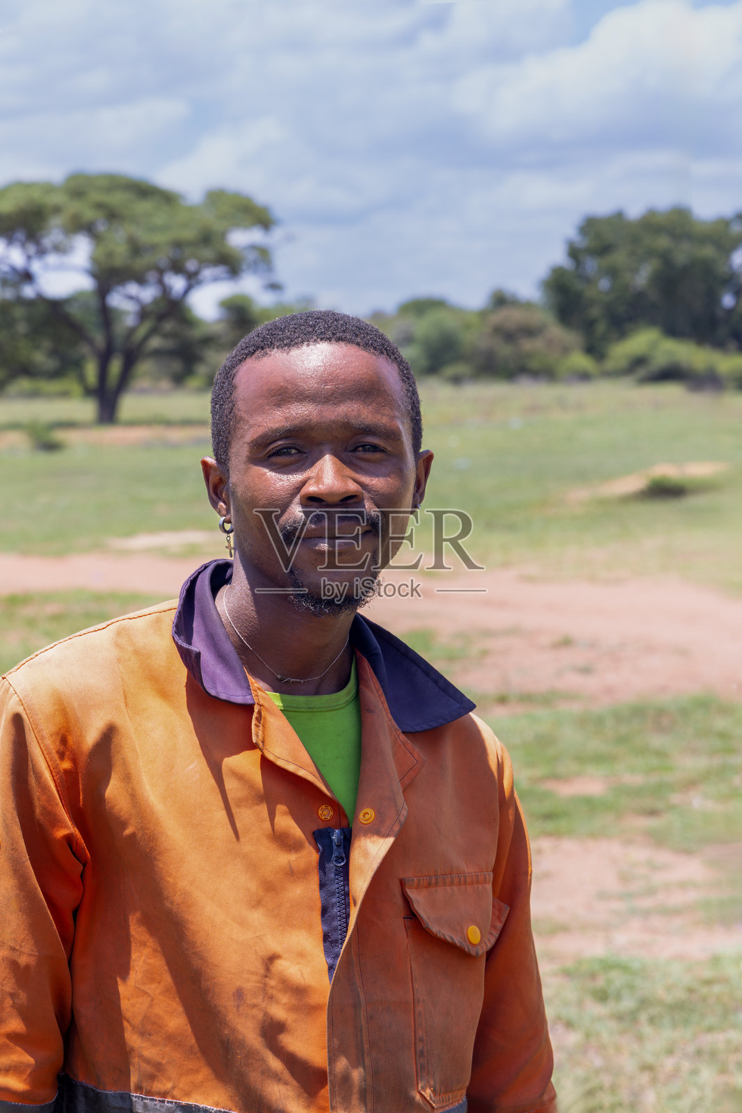 穿着橙色工装的非洲年轻村庄工人，背景是绿色的田野和金合欢树照片摄影图片