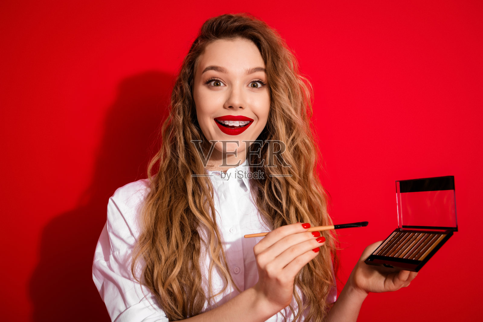一位年轻女子兴奋地拿着化妆刷，在鲜艳的红色背景上展示美妆产品，用于时尚广告宣传。照片摄影图片