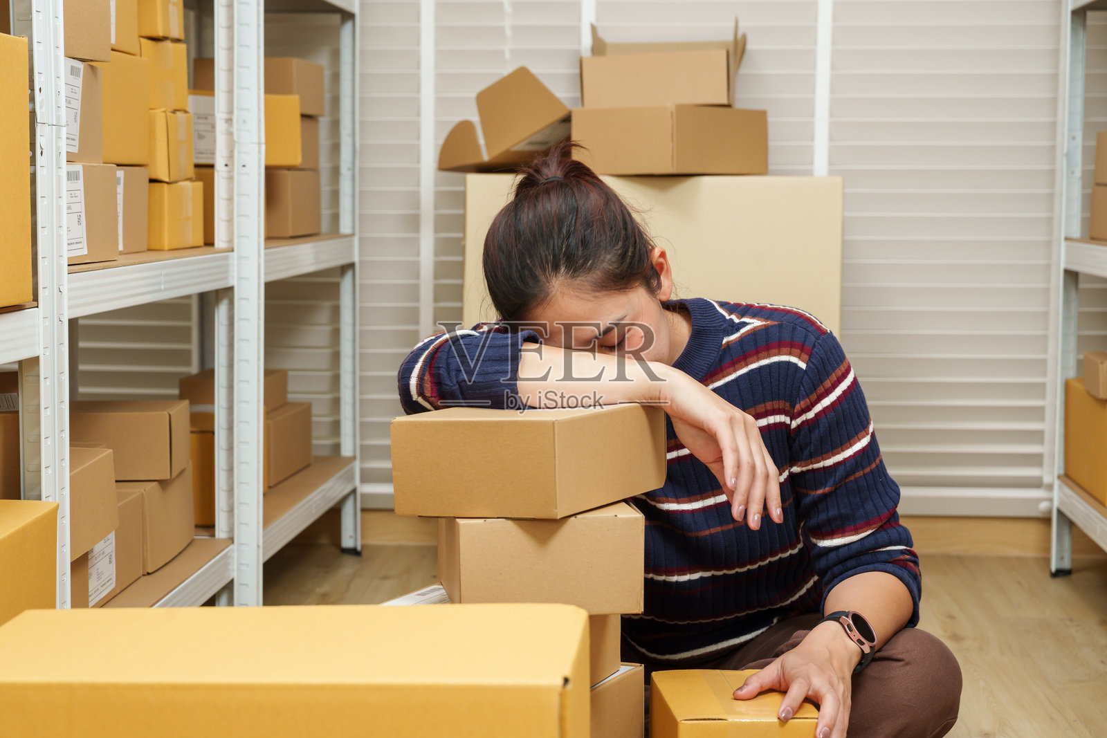 亚洲女性小企业主，疲惫不堪地将头靠在退回的包裹箱上，显示出因电子商务订单问题而在家中仓库里感到的疲惫和沮丧。照片摄影图片