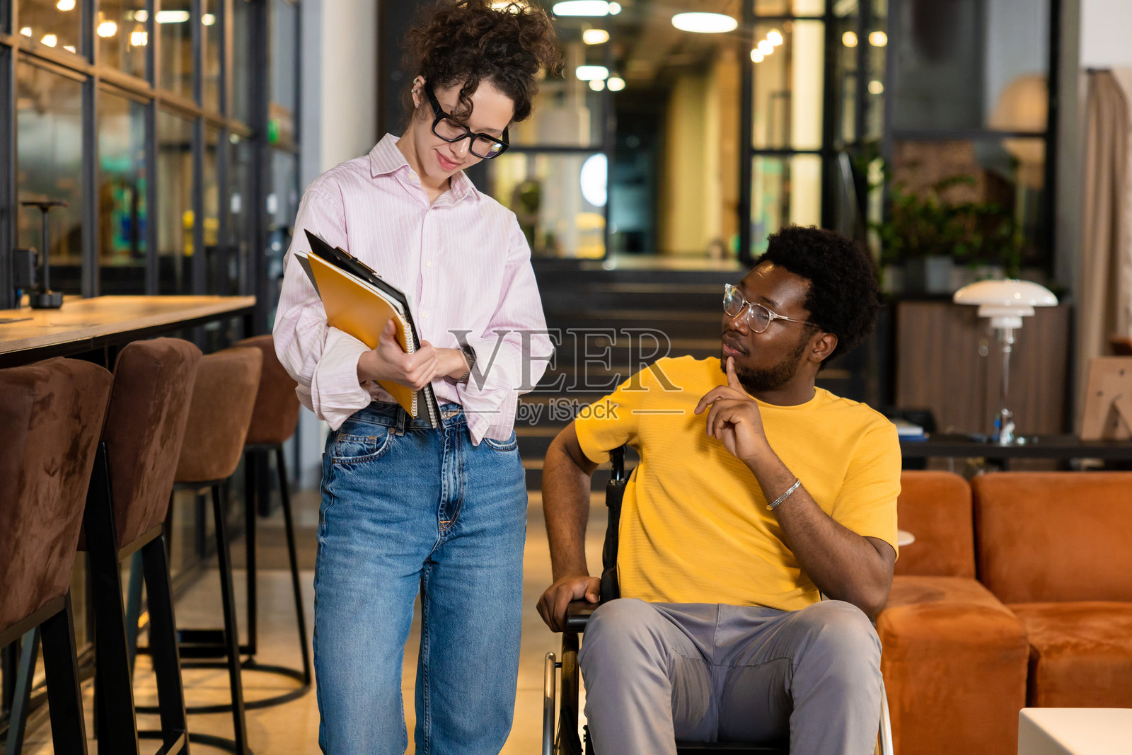年轻女子拿着文件夹，与坐在轮椅上的男子在共享办公室内交谈并摆姿势照片摄影图片