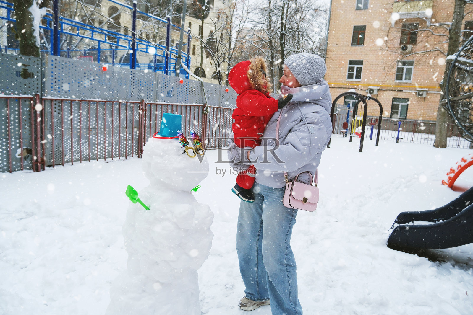 一个欢乐的冬日景象，一位母亲抱着孩子，两人都微笑着看着一个创意装饰的雪人。雪花为公园营造出节日般的喜庆气氛。照片摄影图片