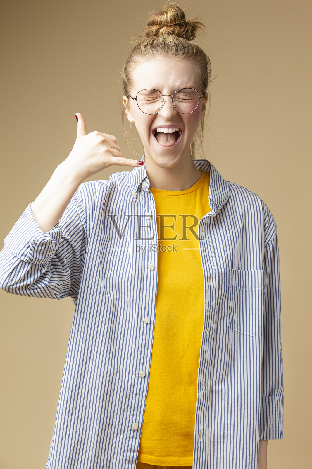 穿着条纹衬衫和眼镜的可爱金发美女，俏皮地用手指比划着想象中的手机，表情滑稽。照片摄影图片