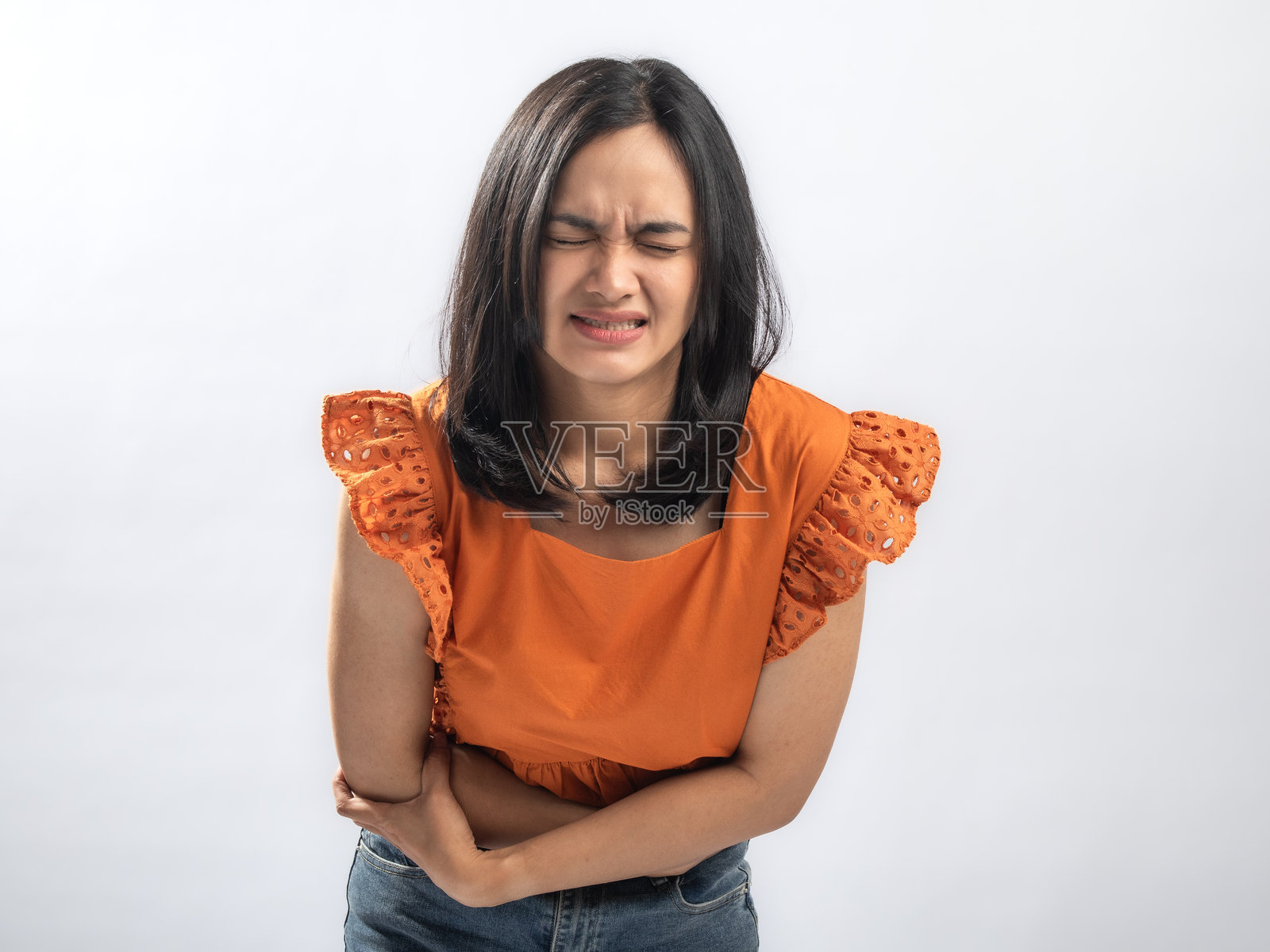 一位身穿橘色荷叶边衬衫的亚洲女性，双手捂着侧腹，表情痛苦地龇牙咧嘴，在影棚的纯白背景下，表现出不适或胃痛照片摄影图片