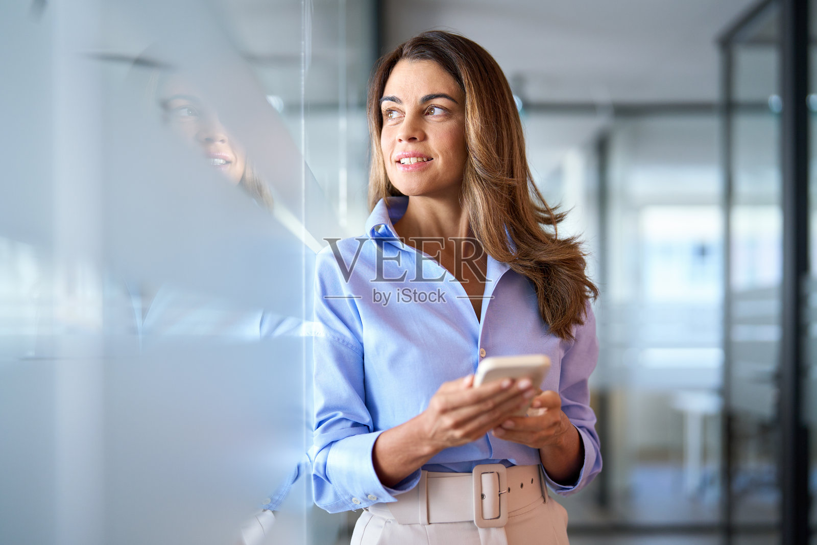 一位成熟的女商人投资者正在办公室站着使用手机，目光望向别处。照片摄影图片