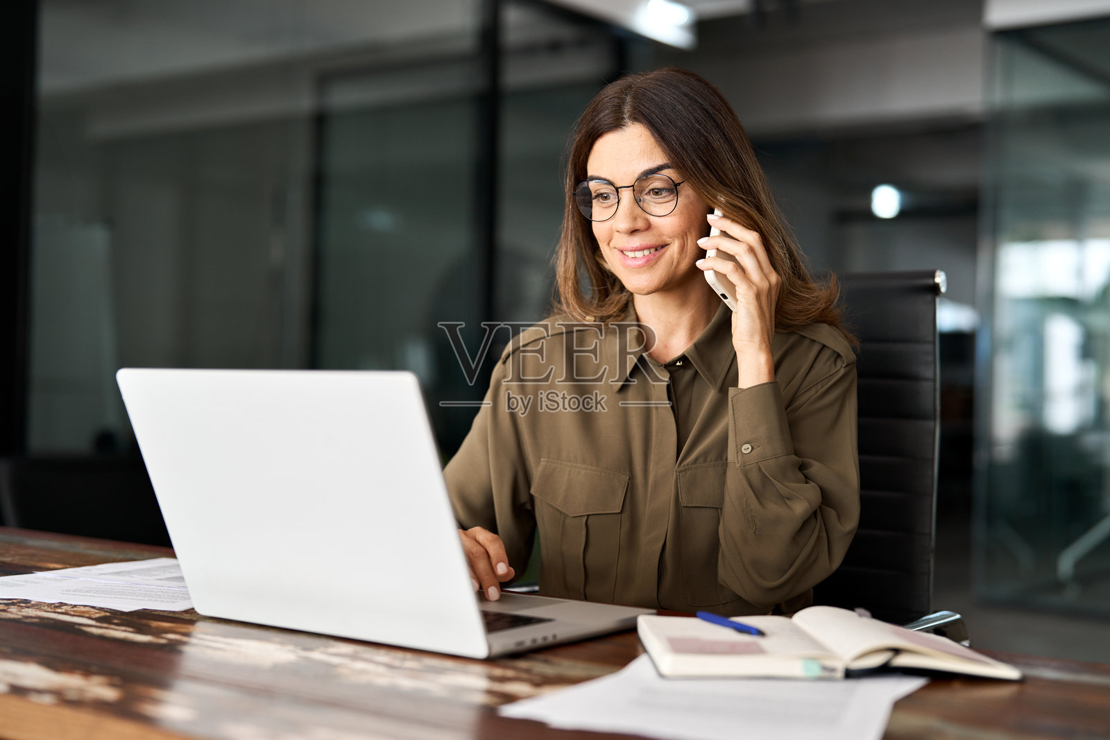 快乐微笑的中年女商人，忙碌的专业人力资源或银行经理，在办公室使用笔记本电脑，通过手机进行商务通话。照片摄影图片
