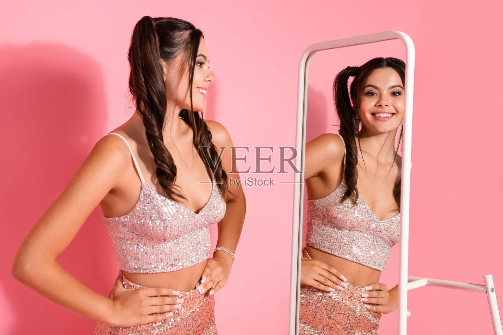 年轻女模特身穿粉色亮片上衣和裙子，在一个俏皮迷人的时尚生活场景中，手持立镜摆拍，适合派对风格的图库摄影。照片摄影图片