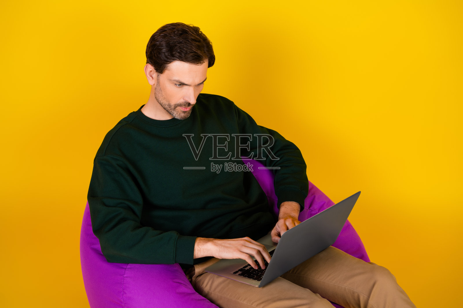 身穿休闲时尚服装的年轻男子坐在紫色椅子上，在明亮的黄色背景前使用笔记本电脑，展示现代工作和科技应用场景。照片摄影图片