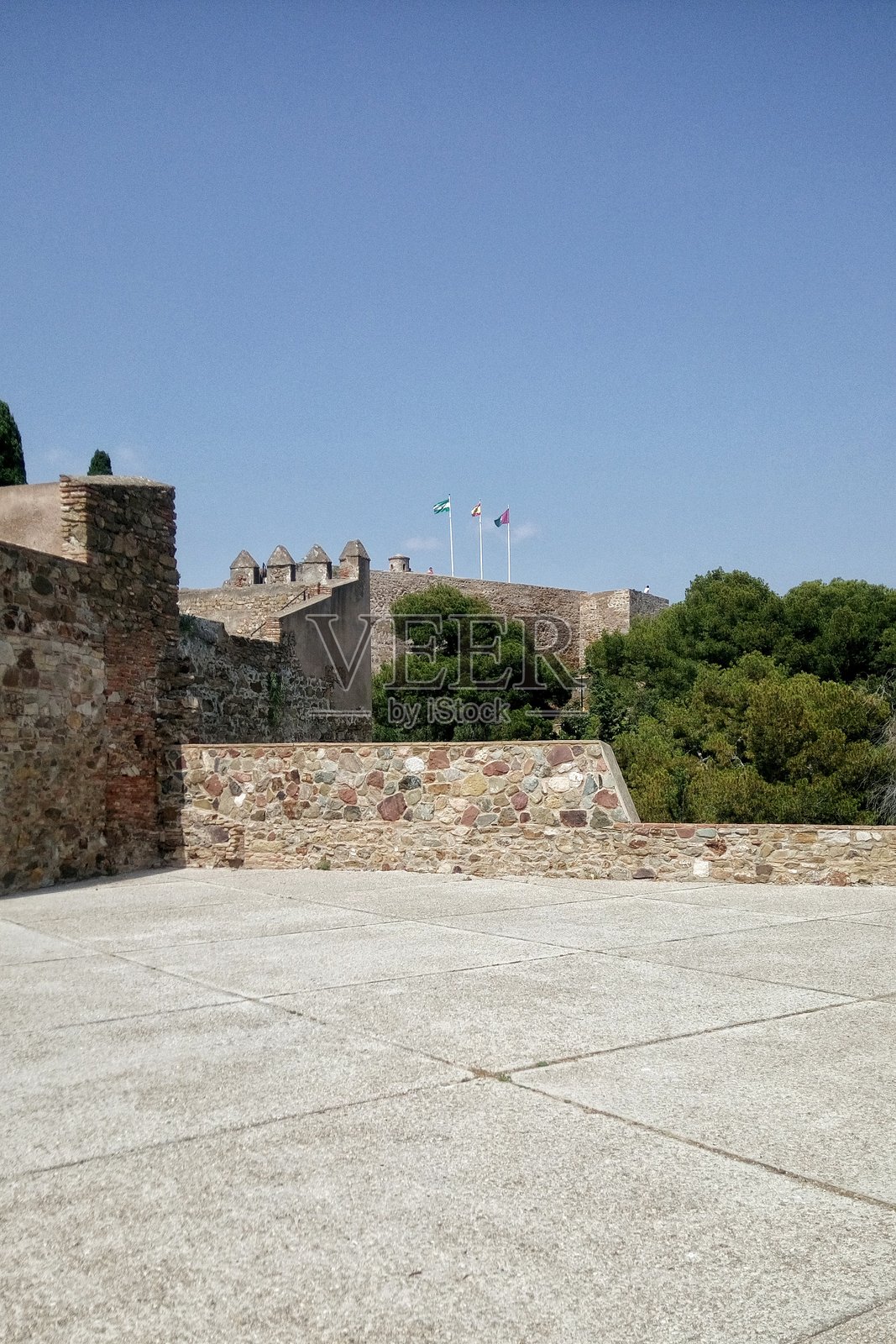 晴朗的蓝天下，西班牙马拉加吉布拉法罗城堡（Gibralfaro Castle）的古老城墙的外部景观，其特色为石砌建筑、混凝土观景台、绿树和旗帜。照片摄影图片