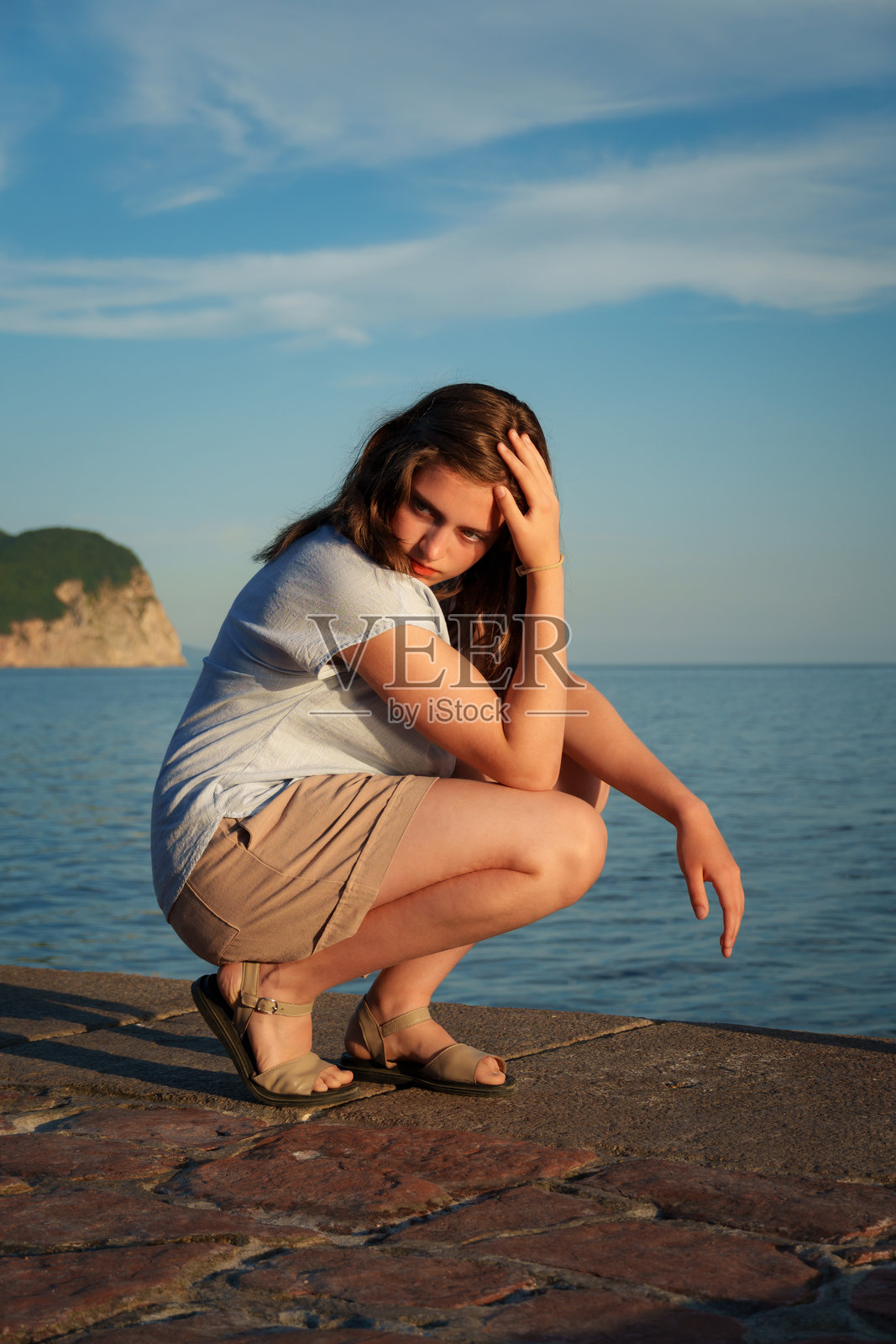 年轻女子在佩特罗瓦茨海边蹲着，手撩头发，看着镜头。中景，背景是海洋和悬崖，夏日夕阳的光线。照片摄影图片