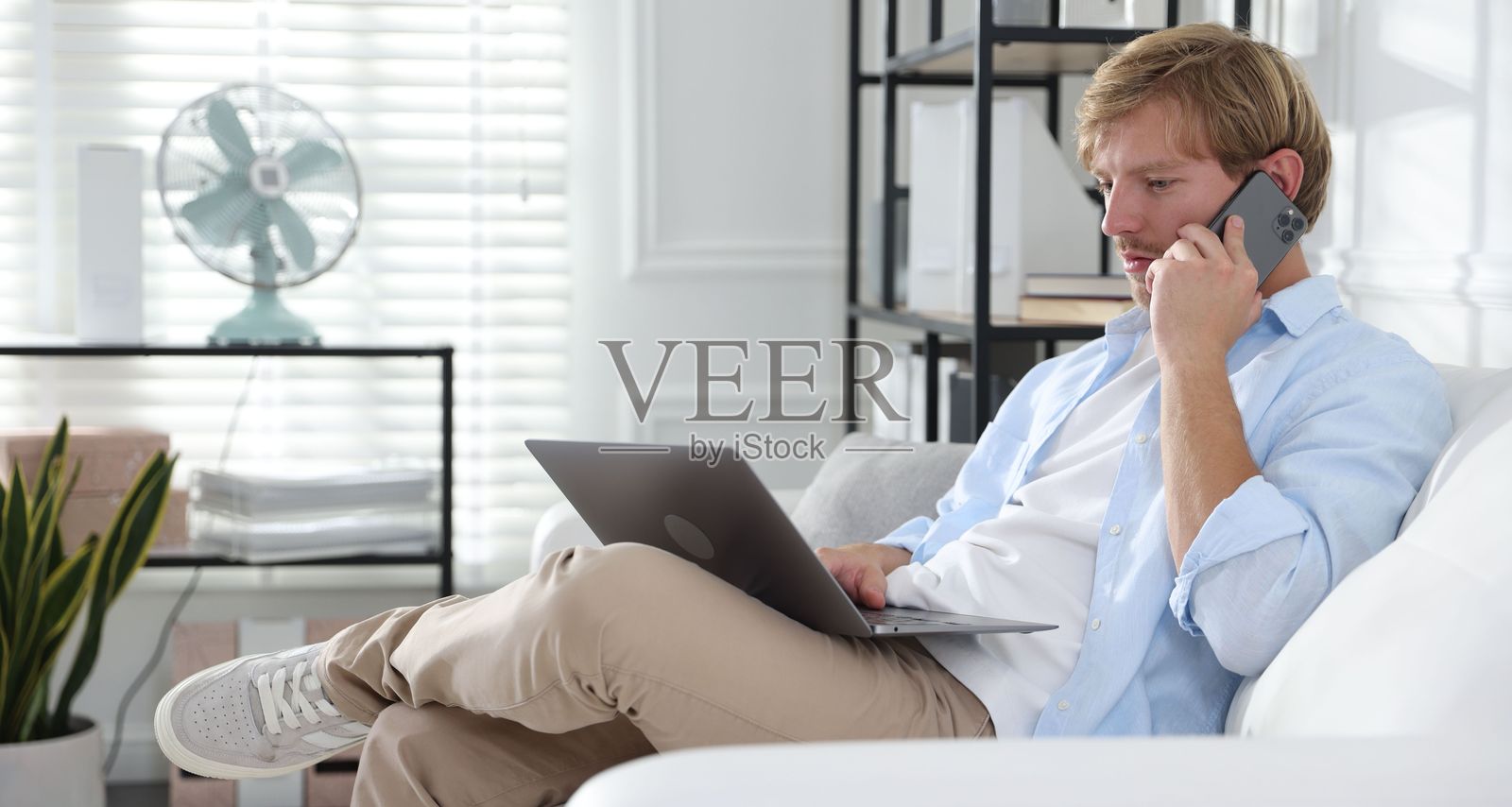 男子在沙发上边用笔记本电脑边打电话照片摄影图片