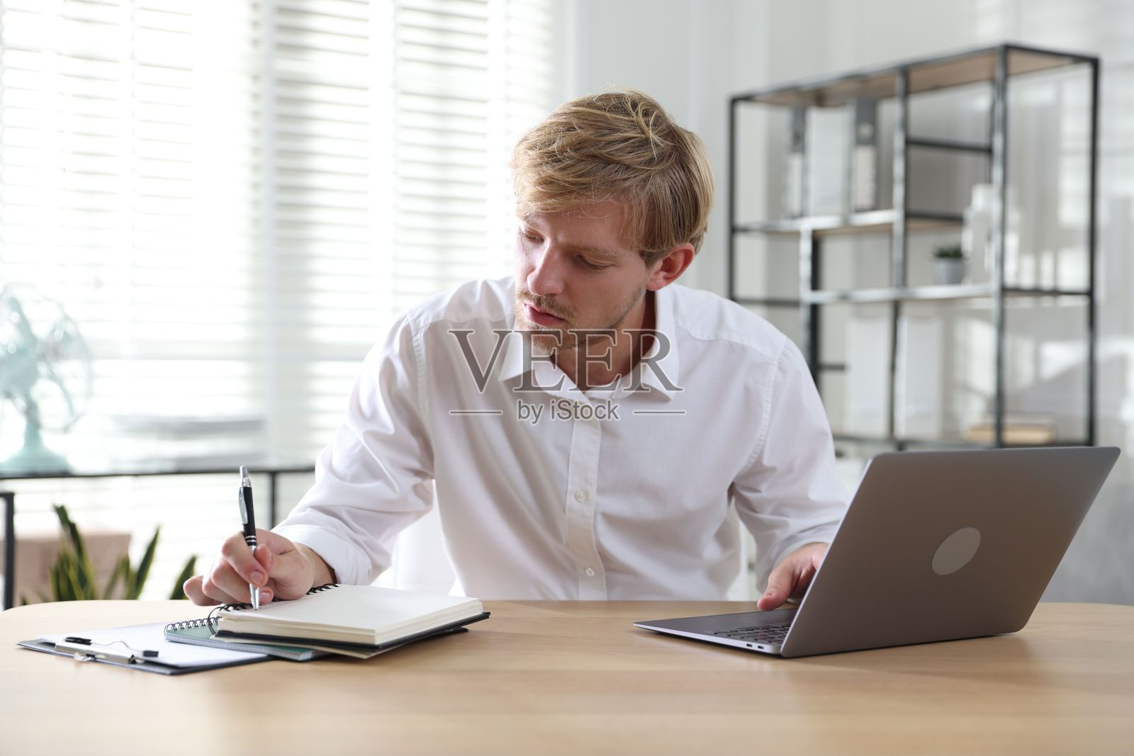 男子在室内木桌旁使用笔记本电脑并做笔记照片摄影图片