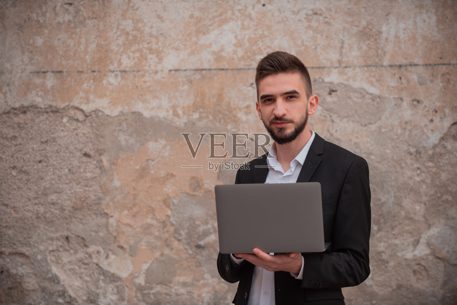 一位年轻快乐的男商人在墙壁背景前使用笔记本电脑。照片摄影图片