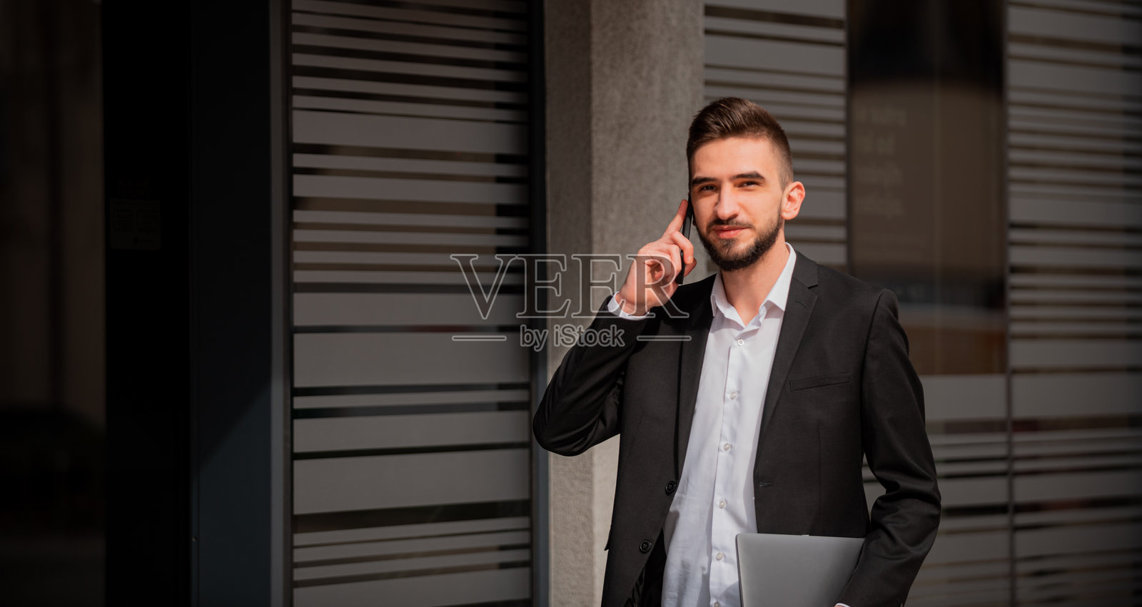 一位年轻快乐的商务男士在办公楼附近户外用手机交谈。照片摄影图片