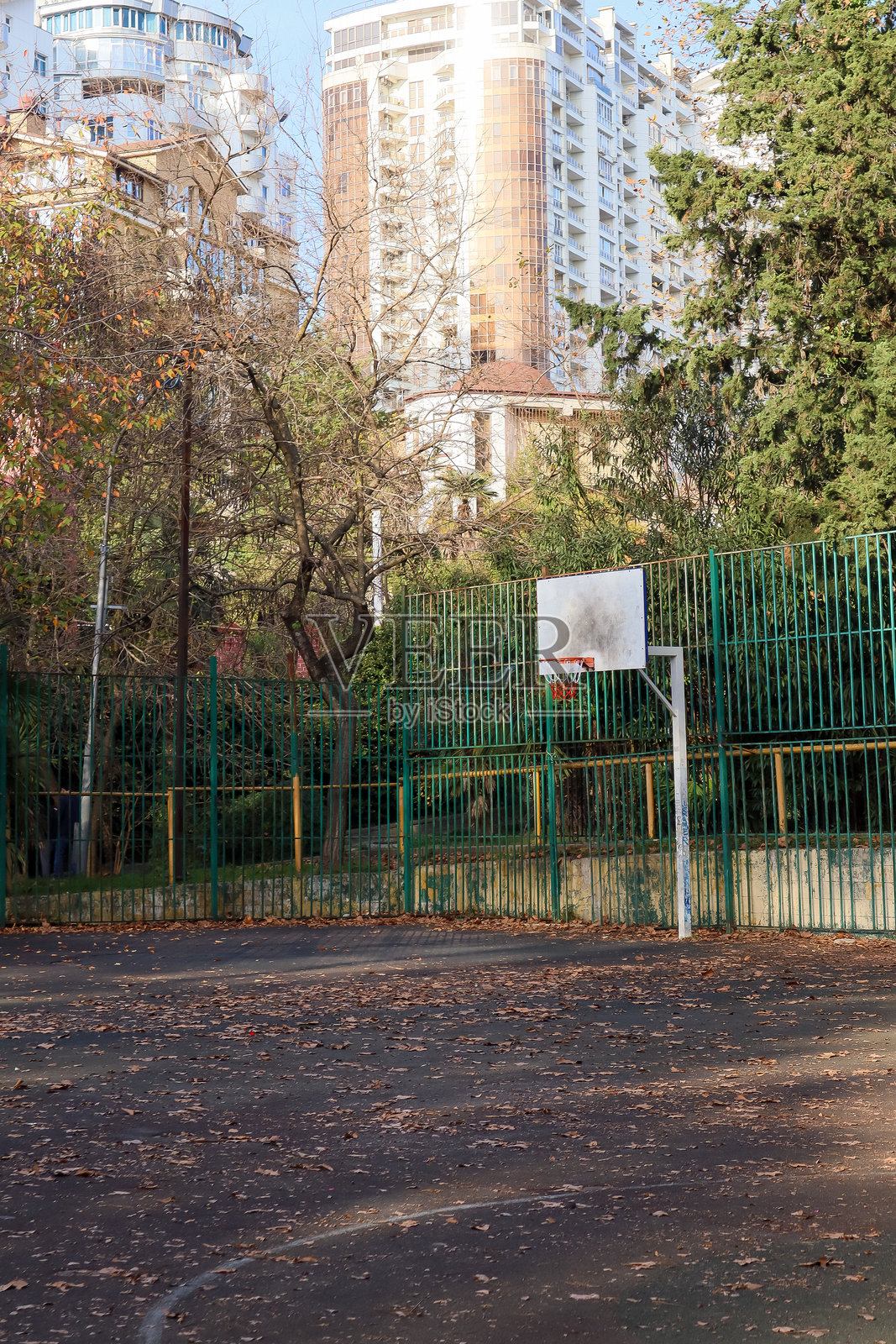 秋日背景下，带围栏的户外篮球场上的篮球架，后面是秋季树木和高楼。照片摄影图片