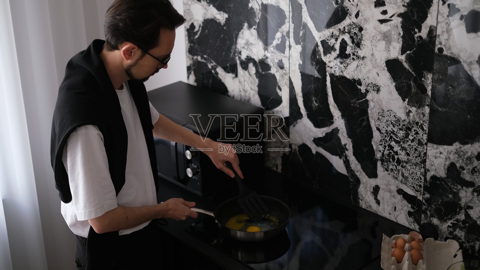 清晨，一位身材苗条的年轻棕发男子正在他现代时尚的厨房里做早餐炒鸡蛋。照片摄影图片