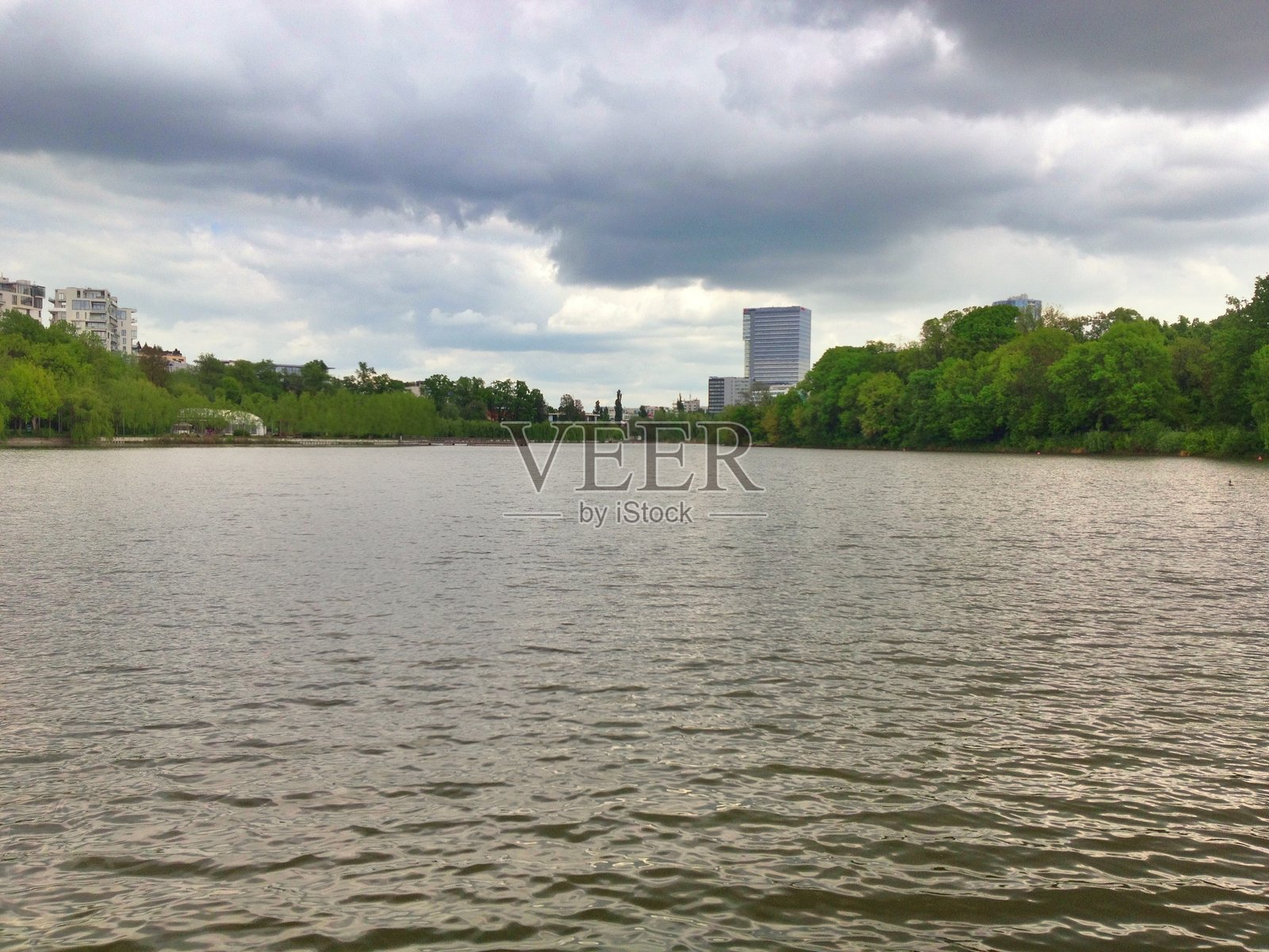 宁静的湖泊，湖边绿树成荫，远方是城市天际线，天空中乌云密布，倒映在水面上照片摄影图片