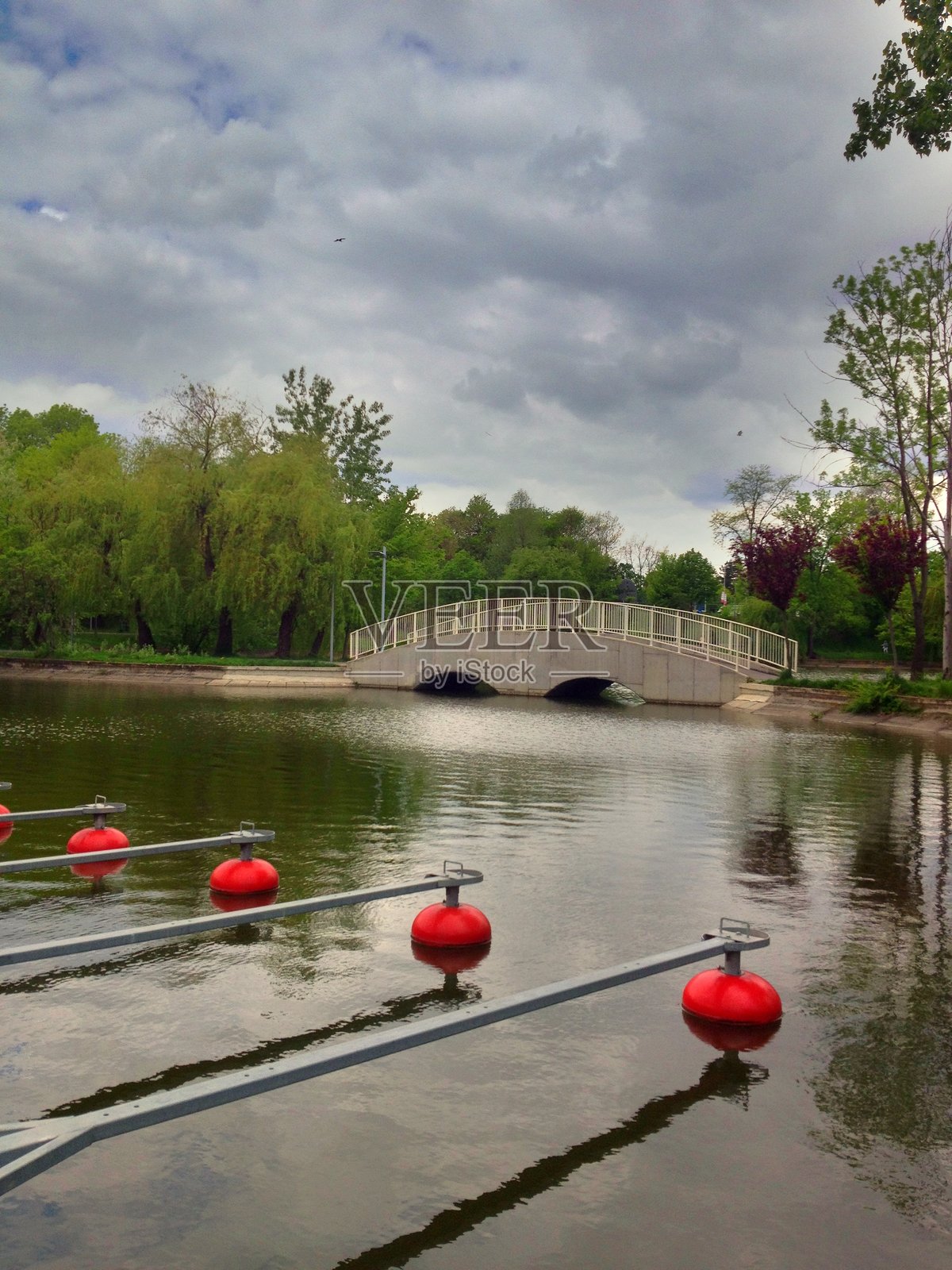 宁静的公园湖景，有白色拱桥、红色浮标和茂密的树木，背景为阴天照片摄影图片