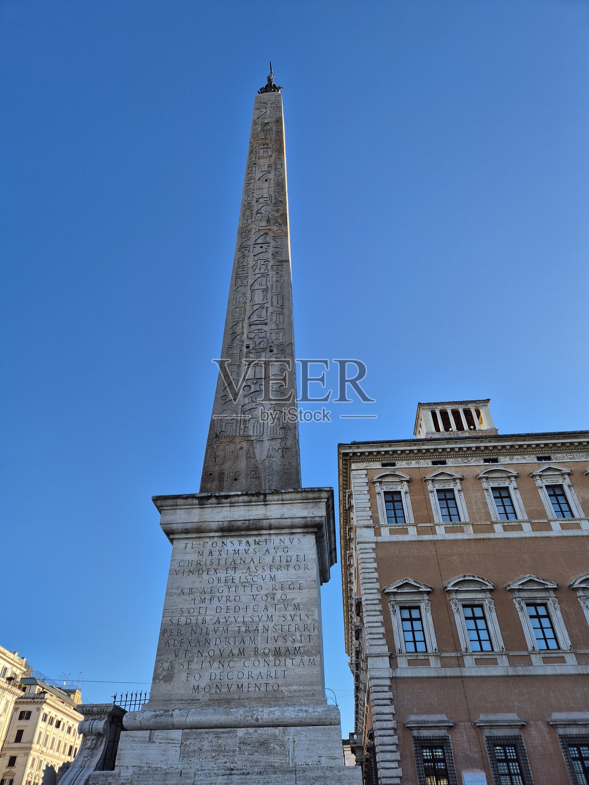 一座高耸的埃及方尖碑，上面刻有象形文字，矗立在刻有拉丁文的石质基座上，位于罗马圣乔万尼拉特兰诺大教堂（Basilica di San Giovanni in Laterano）的正立面旁，背景是晴朗的蓝天。照片摄影图片