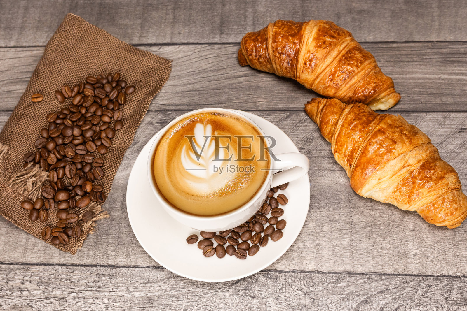 木质背景上的卡布奇诺咖啡，带有花朵拉花、羊角面包和咖啡豆。照片摄影图片