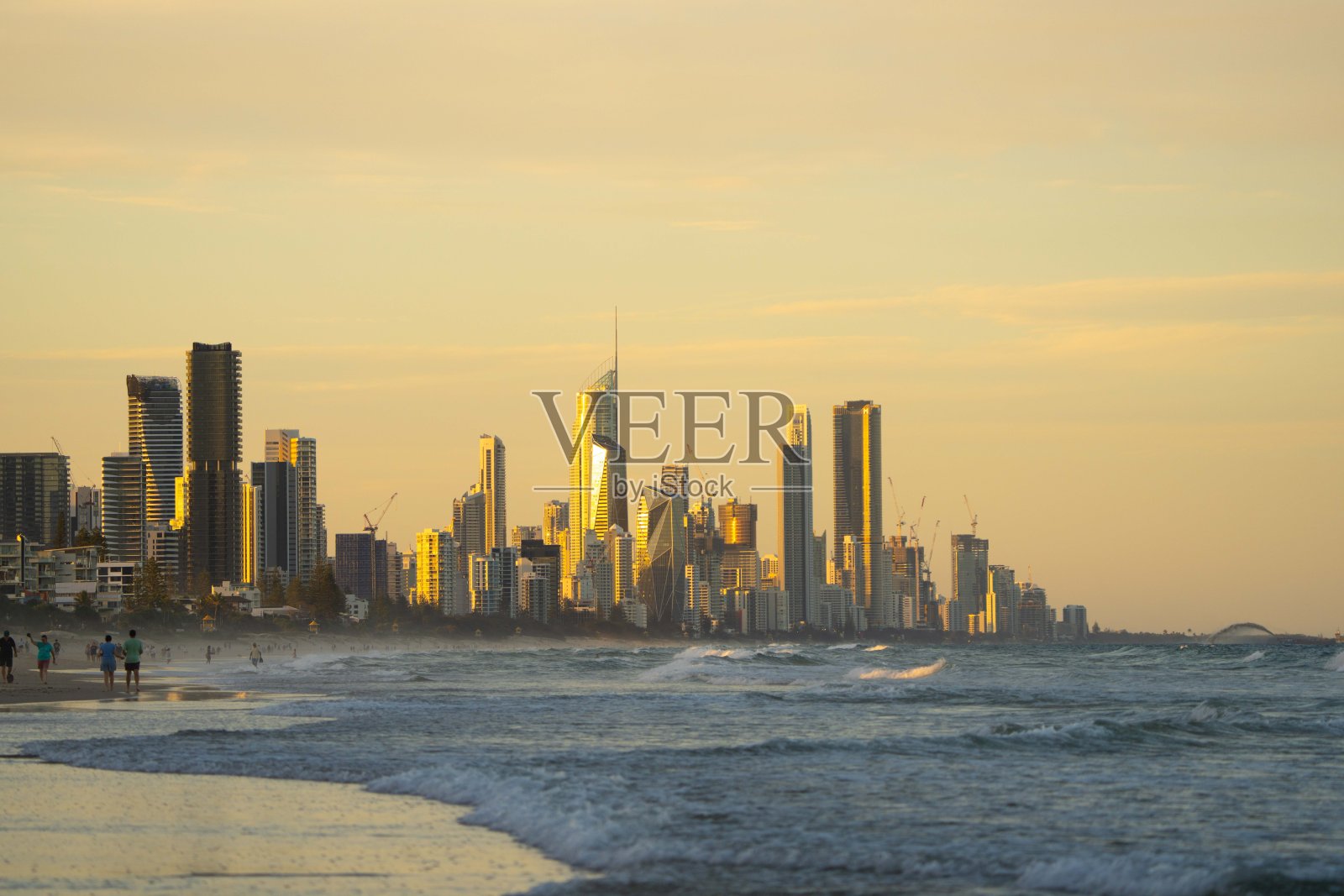 日落时分，蓝色海浪涌向海滩，天空呈黄色，远处可见冲浪者天堂市。照片摄影图片
