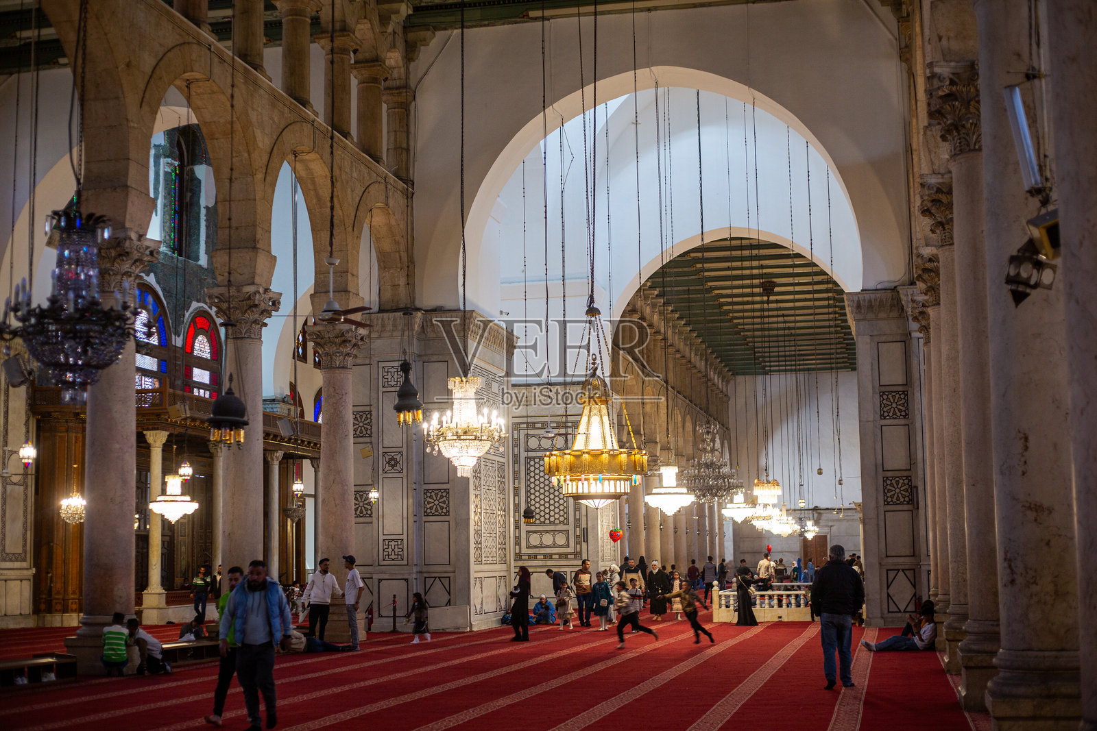 白天,游人们在熙熙攘攘的城市中参观倭马亚清真寺,寺内有拱门、吊灯和红色地毯。照片摄影图片