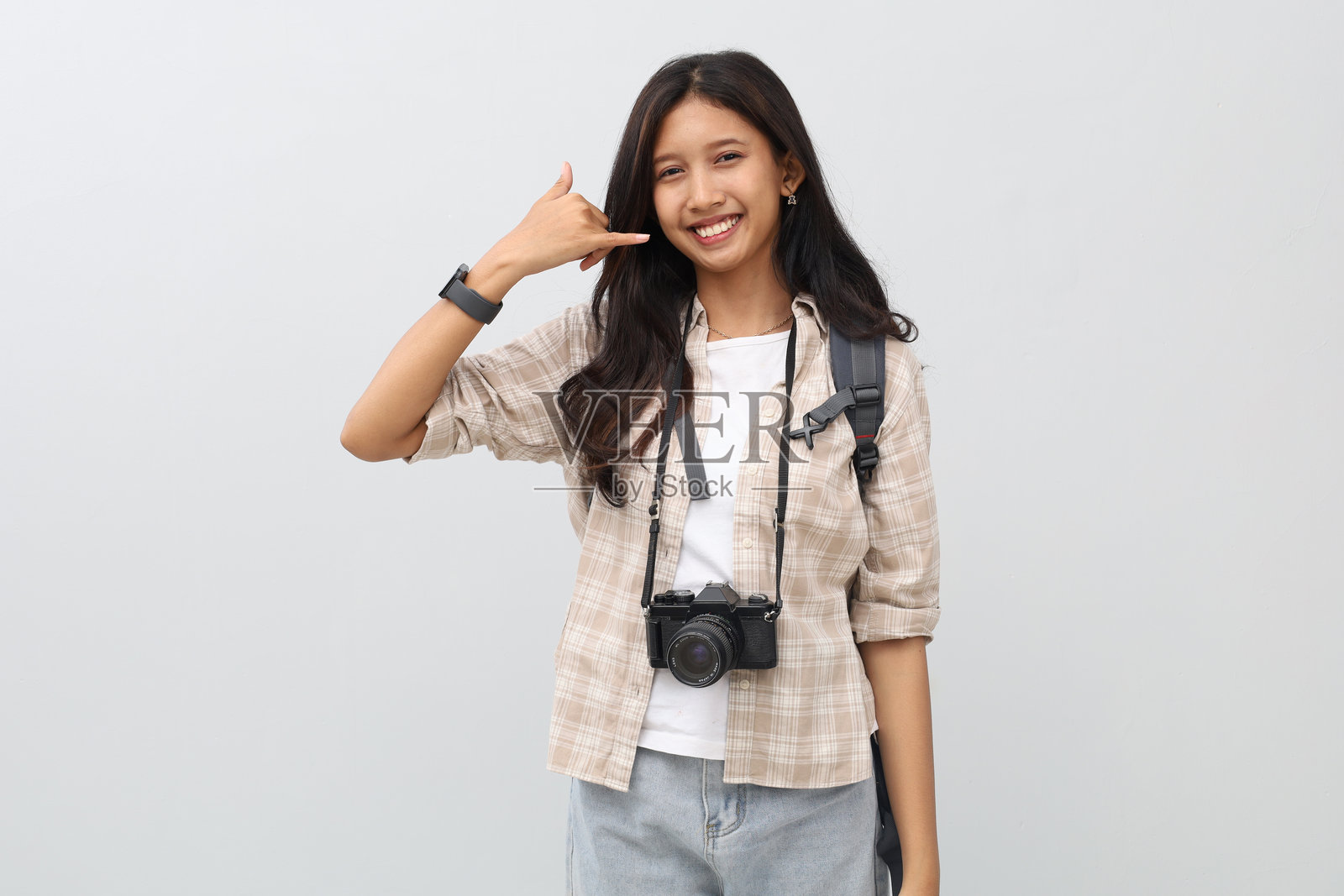 一位年轻的亚洲女性，身穿休闲服，背着背包，脖子上挂着相机，用手指做出打电话的手势，孤立在白色背景上，度假概念。照片摄影图片