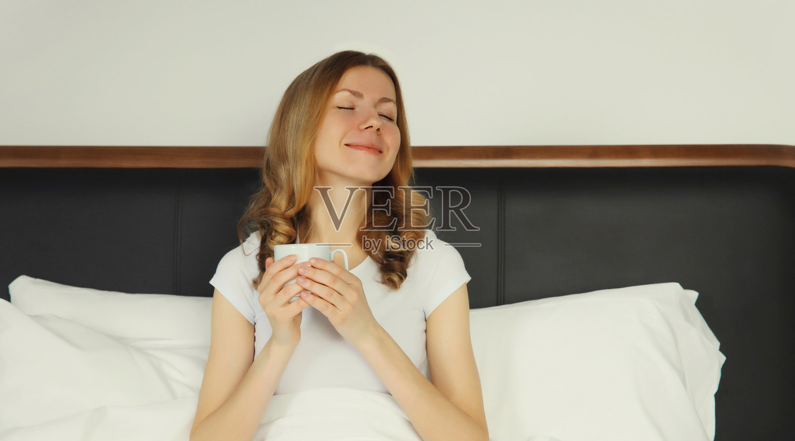 年轻女子在家中床上，面带笑容，享受着一杯热咖啡照片摄影图片