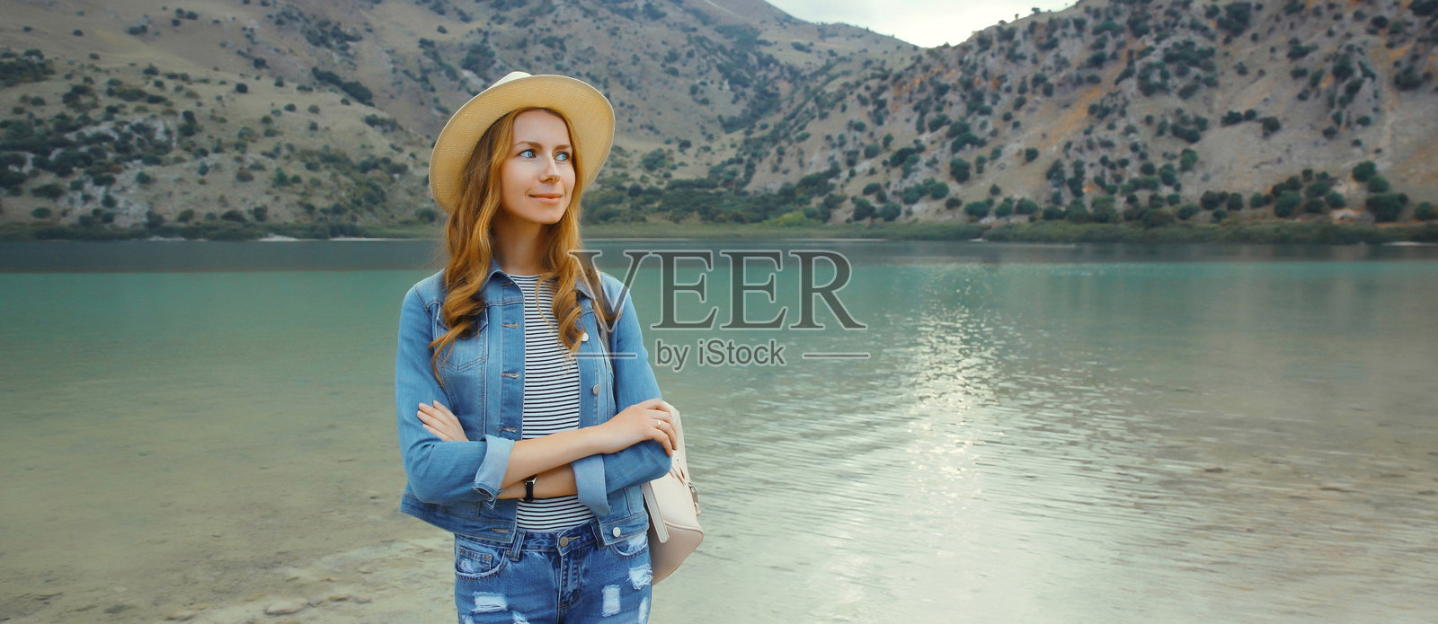 希腊克里特岛，快乐的年轻女游客站在湖边，背景是群山。照片摄影图片